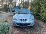 Front view of a silver 2007 Mazda 3 hatchback parked in a wooded area, showing its distinctive grille and headlights.