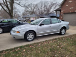 2003 Oldsmobile Alero GL in silver, parked on a driveway with a grassy area, showcasing alloy wheels.