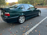 1997 BMW E36 M3 in green with grey interior, modified features and OEM Style 24 wheels parked on leaf-covered street.
