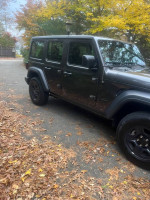 2023 Jeep Wrangler Sport 4WD parked on a leaf-covered driveway, showcasing its grey exterior and rugged design.