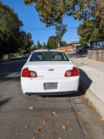 Rear view of a 2012 Chevrolet Malibu LS in white, parked on a street with some wear and tear visible.