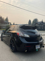 2010 Mazdaspeed3 black hatchback with custom modifications parked on the street, showcasing upgraded wheels and exhaust.