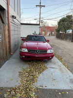 Red 1997 Mercedes-Benz SL500 with peeling paint and leather tears parked on a driveway surrounded by leaves.