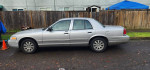 2007 Ford Crown Victoria silver sedan parked on the street with wooden fence in background.
